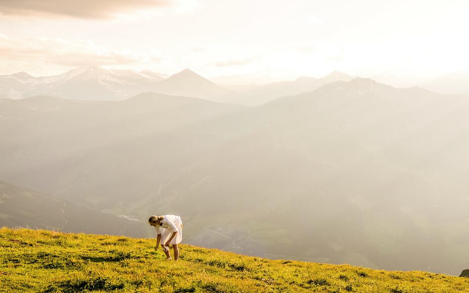 A person in a white dress bends down on a golden alpine meadow, with soft mountain silhouettes in warm sunlight in the background.