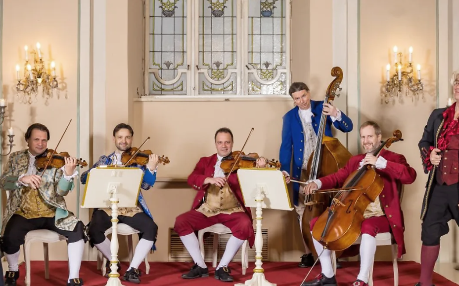 Baroque music ensemble in historical costumes, six musicians with instruments in an elegant hall with chandeliers and stained glass windows.