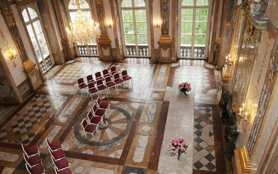 Magnificent baroque hall with checkered marble floor, golden chandeliers, and red velvet chairs, large windows overlooking greenery.