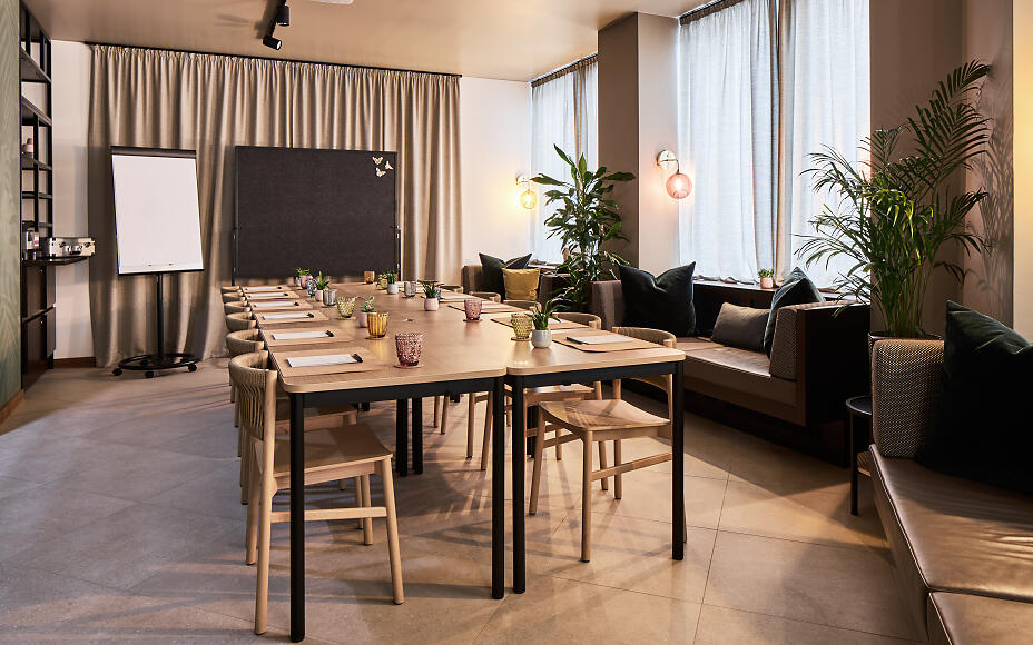 Moderner Meetingraum mit holzfarbenen Tischen, grauen Stühlen und grünen Zimmerpflanzen in elegantem Design.