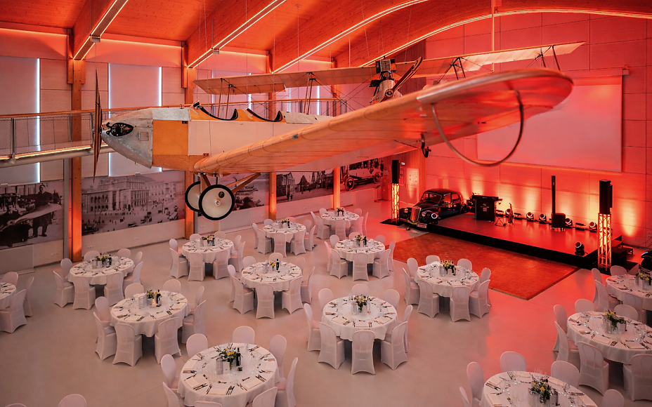 Die außergewöhnliche Eventlocation im fahr(T)raum Mattsee vereint historische Luftfahrt mit stilvoller Veranstaltungsatmosphäre. Unter einem imposanten historischen Flugzeug erstreckt sich ein elegant gedeckter Saal mit runden Tischen, Bühne und eindrucksvoller roter Lichtinszenierung – ideal für Galas, Firmenfeiern, Dinner-Events und exklusive Abendveranstaltungen.