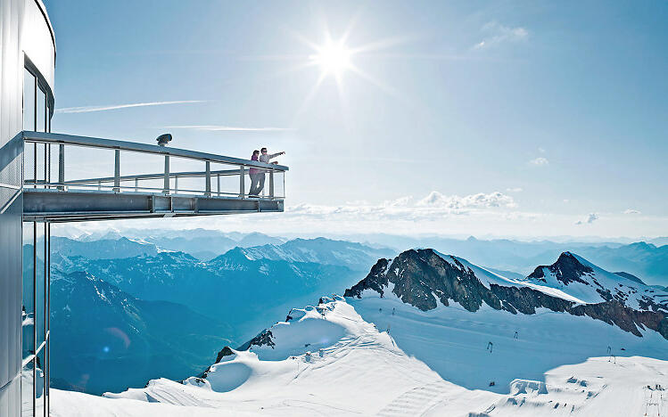 Moderne Aussichtsplattform am Kitzsteinhorn mit Blick auf verschneite Alpenberge unter strahlend blauem Himmel und Sonnenschein.