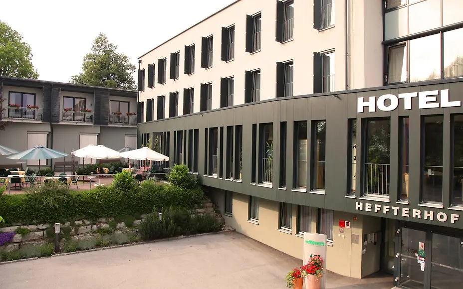 The modern hotel building combines clean lines with warm green and natural stone. Large window fronts and flower-adorned balconies give the house a welcoming charm. A terrace with sunshades in the courtyard invites guests to relax outdoors.