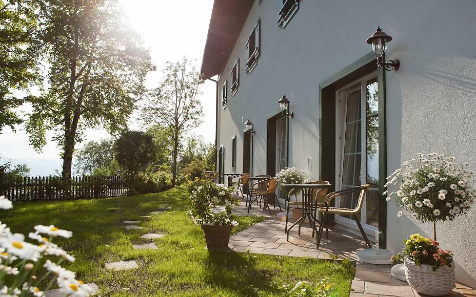 Sonnendurchflutete Terrasse eines weißen Landhauses mit gemütlichen Gartenmöbeln, umgeben von blühenden Gänseblümchen und grünem Rasen.