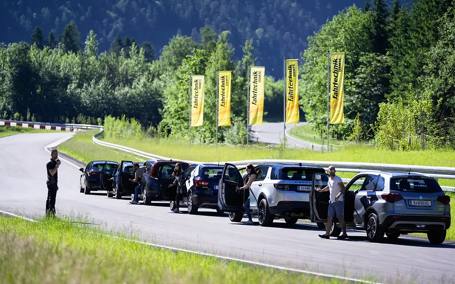 Eine Reihe moderner Fahrzeuge hält auf der kurvenreichen Strecke des ÖAMTC Fahrtechnik Zentrums Saalfelden. Teilnehmer steigen aus und bereiten sich auf das Fahrtraining vor, während gelbe Fahnen entlang der Strecke wehen und die grüne alpine Landschaft eine eindrucksvolle Kulisse für das Sicherheitstraining bildet.