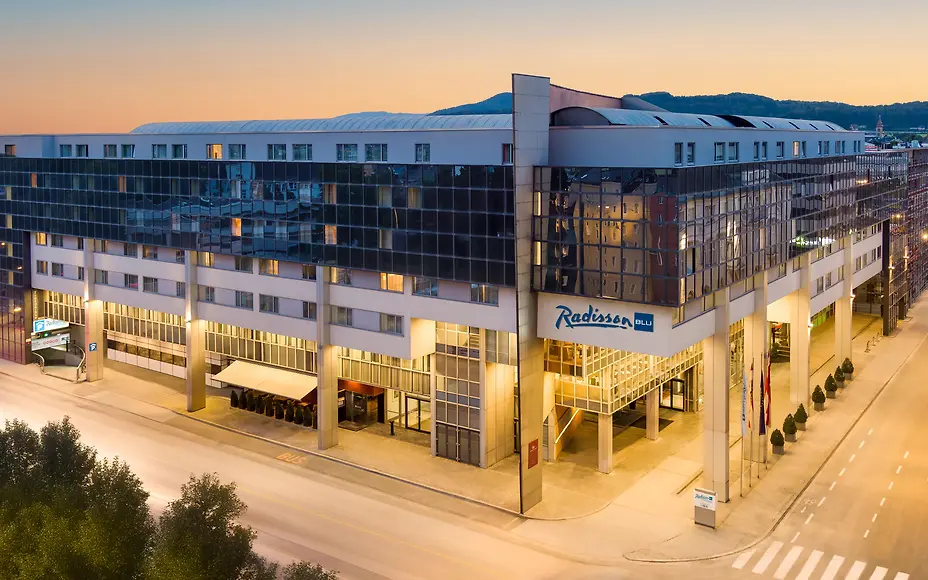 Das moderne Radisson Blu Hotel erhebt sich in klarer Architektur entlang einer ruhigen Straße bei Abenddämmerung. Großzügige Glasfassaden spiegeln das warme Licht des Sonnenuntergangs, während der einladende Eingangsbereich und dezente Beleuchtung urbanen Komfort und Exklusivität vermitteln.