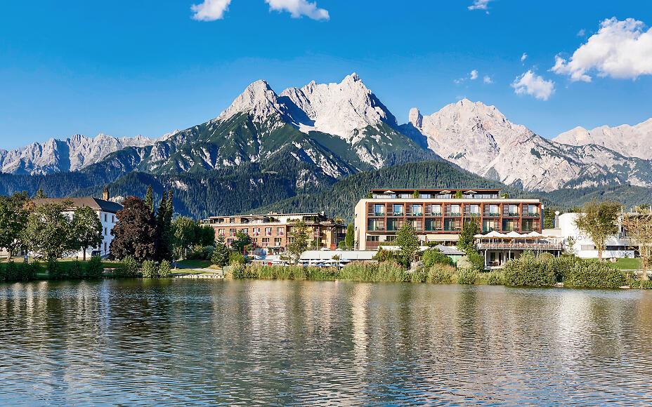 Modern hotel by a lake shore with majestic Alpine mountains in the background, surrounded by lush vegetation and reflective water surface.