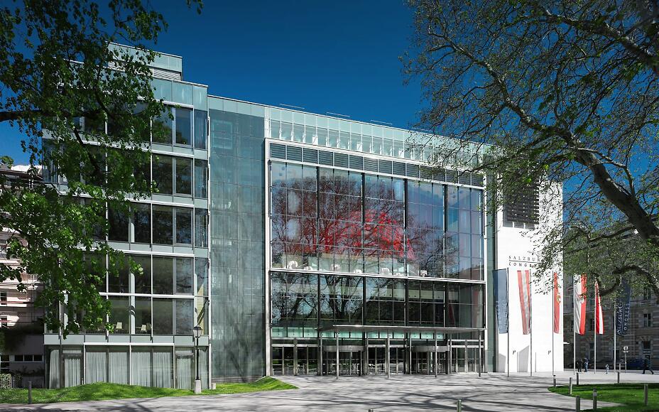 The modern glass building of Salzburg Congress gleams in the sunlight, framed by lush green trees. Its glass façade reflects the blue sky and surrounding nature, while Austrian flags adorn the prestigious main entrance.
