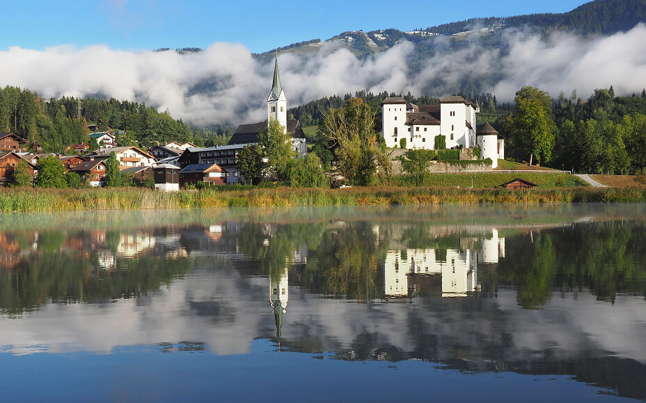 Idyllic lake landscape in Goldegg with reflective lake, white church, and historic buildings against misty mountain backdrop.