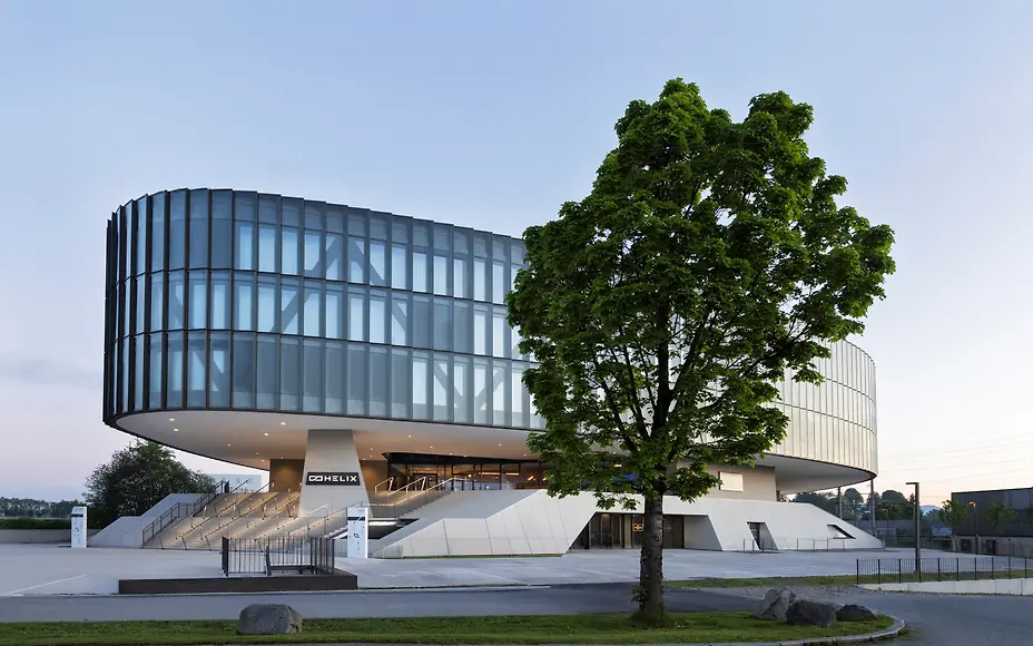 Das futuristische The Passenger Hotel Salzburg beeindruckt mit markanter, ovaler Glasarchitektur und großzügigen Freiflächen. Im Vordergrund setzt ein großer Baum einen natürlichen Kontrast zur modernen Fassade und unterstreicht die elegante, urbane Atmosphäre des Gebäudes.