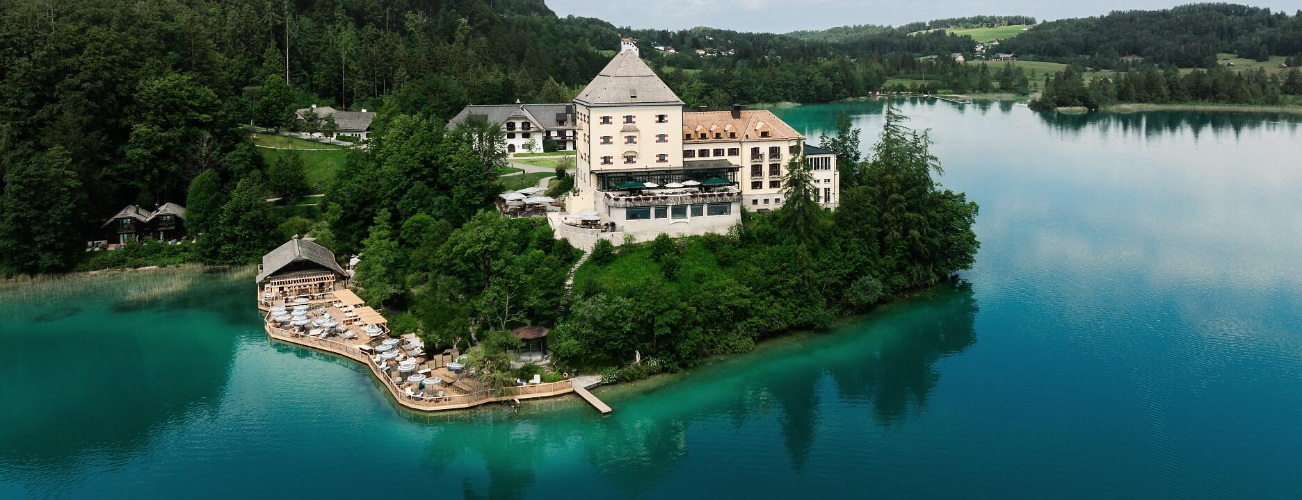 Historisches Schlossgebäude auf einer bewaldeten Halbinsel im türkisfarbenen Fuschlsee, umgeben von grünen Berglandschaften und blauem Himmel.