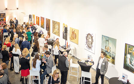 Kunstausstellung mit zahlreichen Besuchern in einem hellen Galerieraum. Verschiedene Kunstwerke hängen an den Wänden, während Menschen in Gruppen stehen und miteinander kommunizieren.